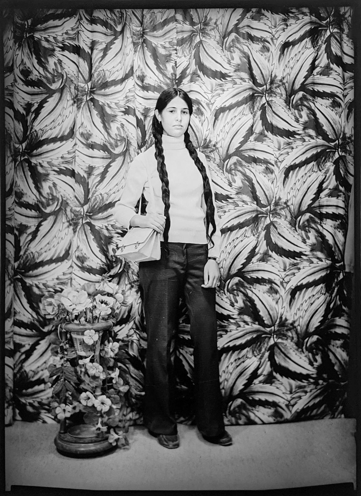 1960s black and white photograph of a woman standing in a white turtleneck and dark pants with her hair in two braids