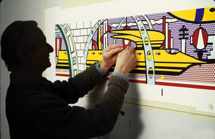 Photograph of a man in dark silhouette working on a blue, red, and yellow painting of a vehicle