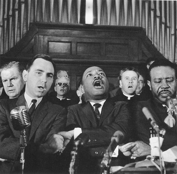 Black and white photograph of Martin Luther King leading the singing of We Shall Overcome after eulogizing a slain civil rights crusader, the Reverend James Reeb, at Brown Chapel in Selma, Alabama