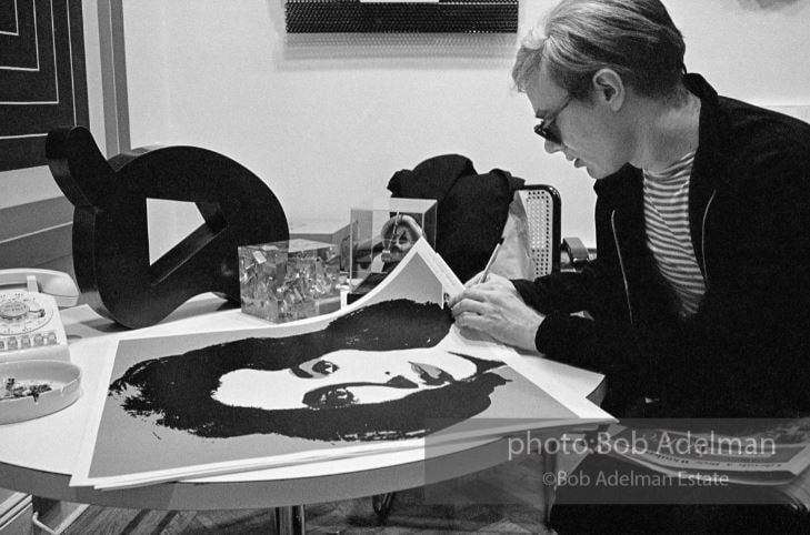 Black and white photograph of Andy Warhol signing Liz Taylor prints at a table in the Leo Castelli Gallery