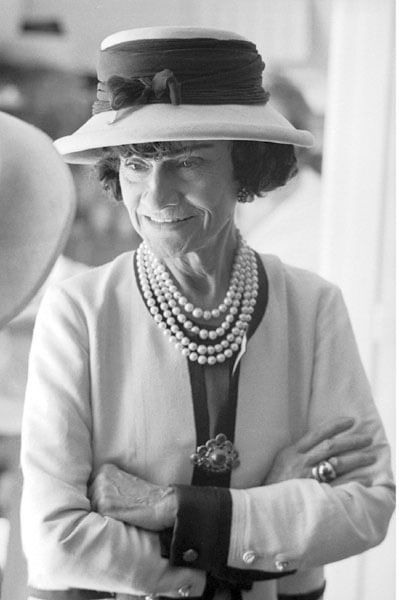 Black and white photograph of Designer Coco Chanel in her private apartment at the House of Chanel, wearing her trademark look of costume jewelry mixed with gems and pearls