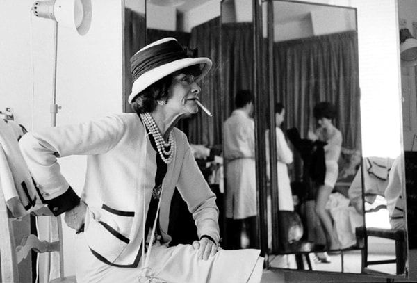 Black and white photograph of Coco Chanel smoking a cigarette with Jean, her right-hand “dresser”, and model Jackie Rogers in the mirror behind her