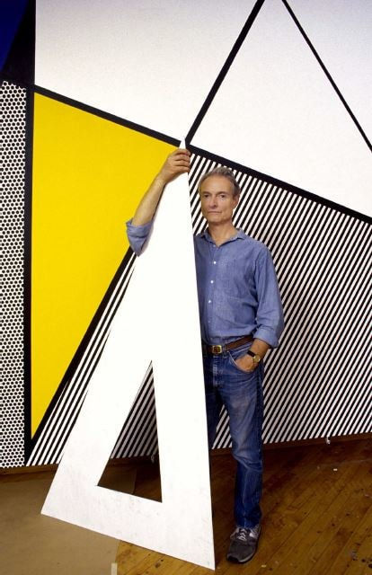 Photograph of Roy Lichtenstein holding a large white isosceles triangular object standing in front of a yellow, black, and white geometric mural