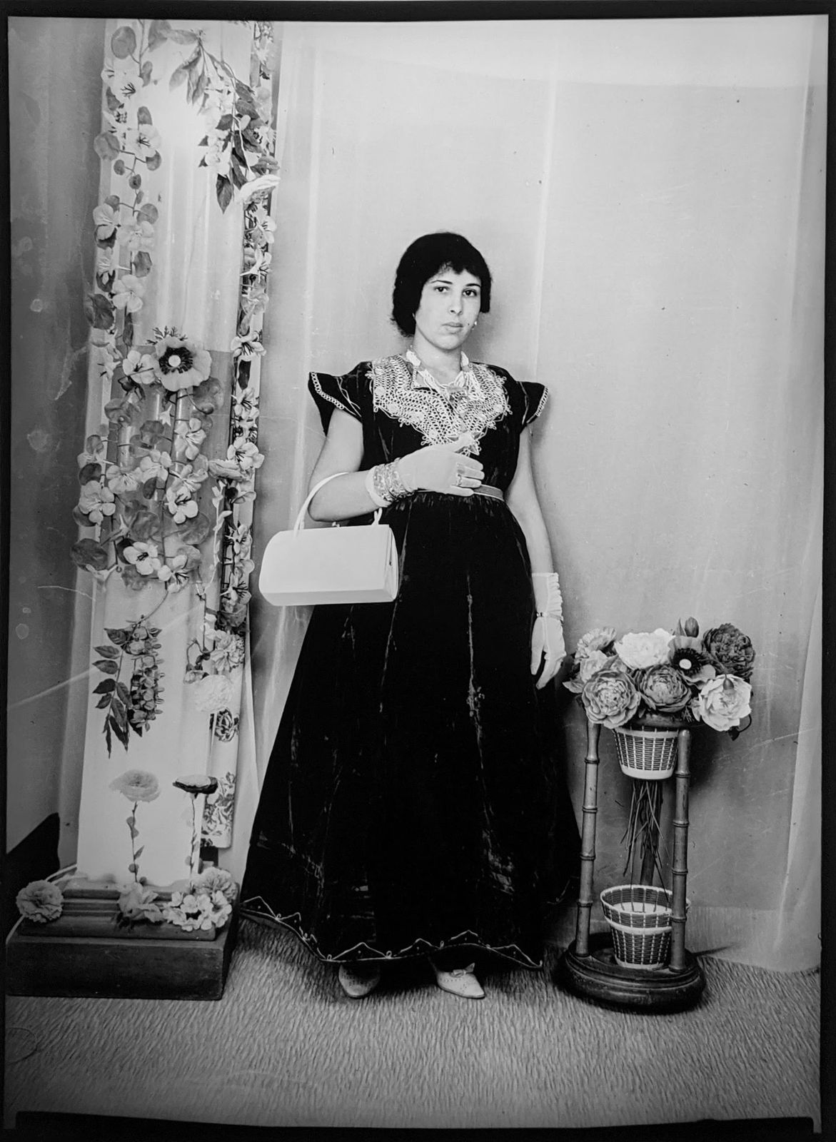 1960s black and white photograph of a woman standing in a black embroidered dress with her left hand to her chest and a white purse resting on her elbow