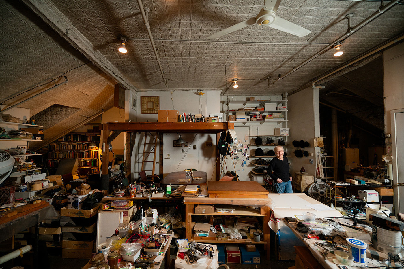 An artist, Gilda Pervin, standing amidst an array of work desks, art materials, and books in a loft setting