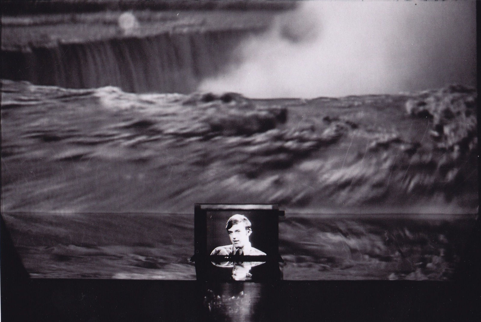A small television screen showing a young man placed on top of a mirror. Behind it is a screen showing videos of Niagara Falls. This video is reflected in the mirror underneath.