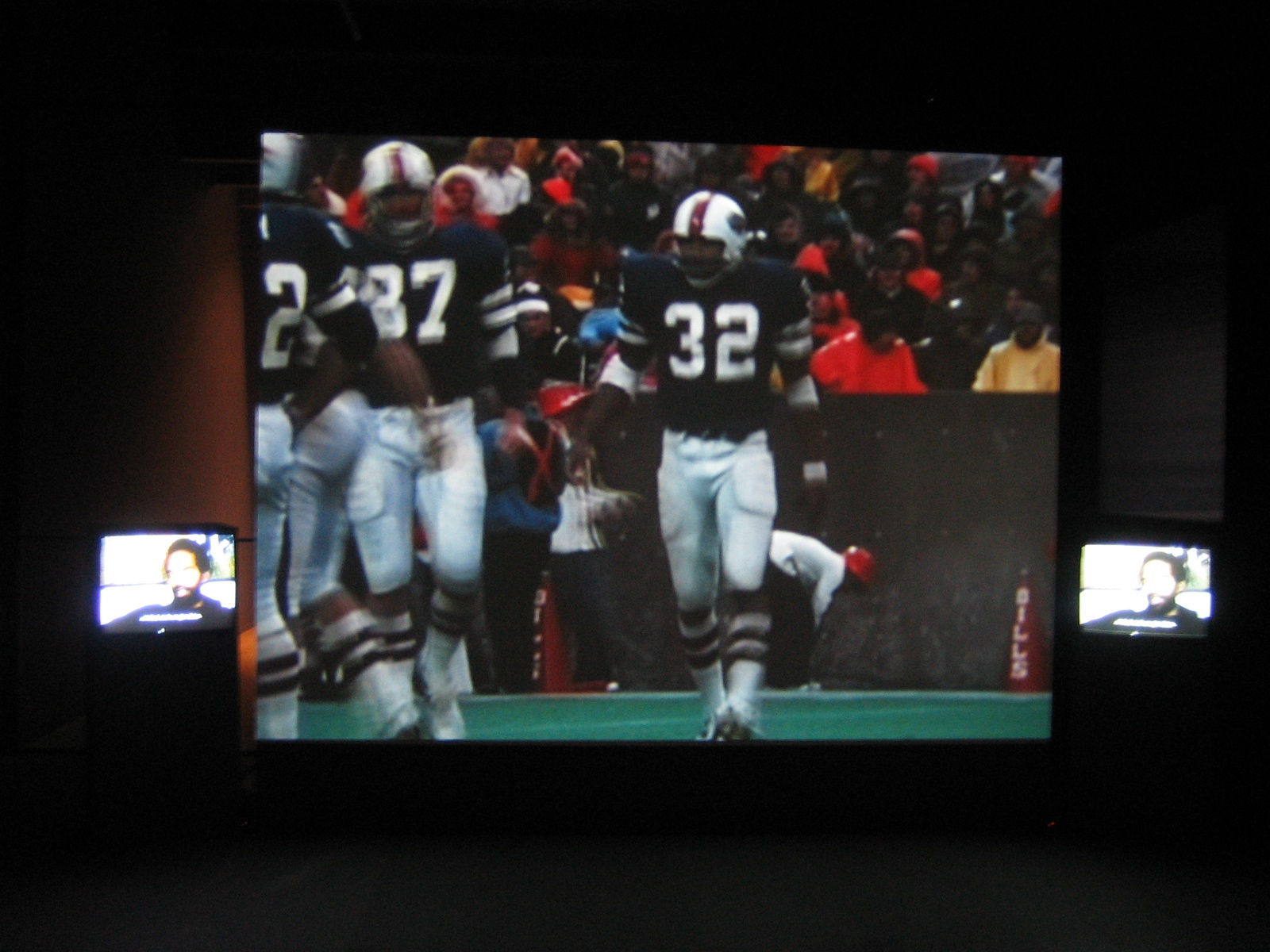 Three screens hanging on a dark wall. The small screens on the outside show a video of a man being interviewed. The large screen in the center shows football players at an NFL game.