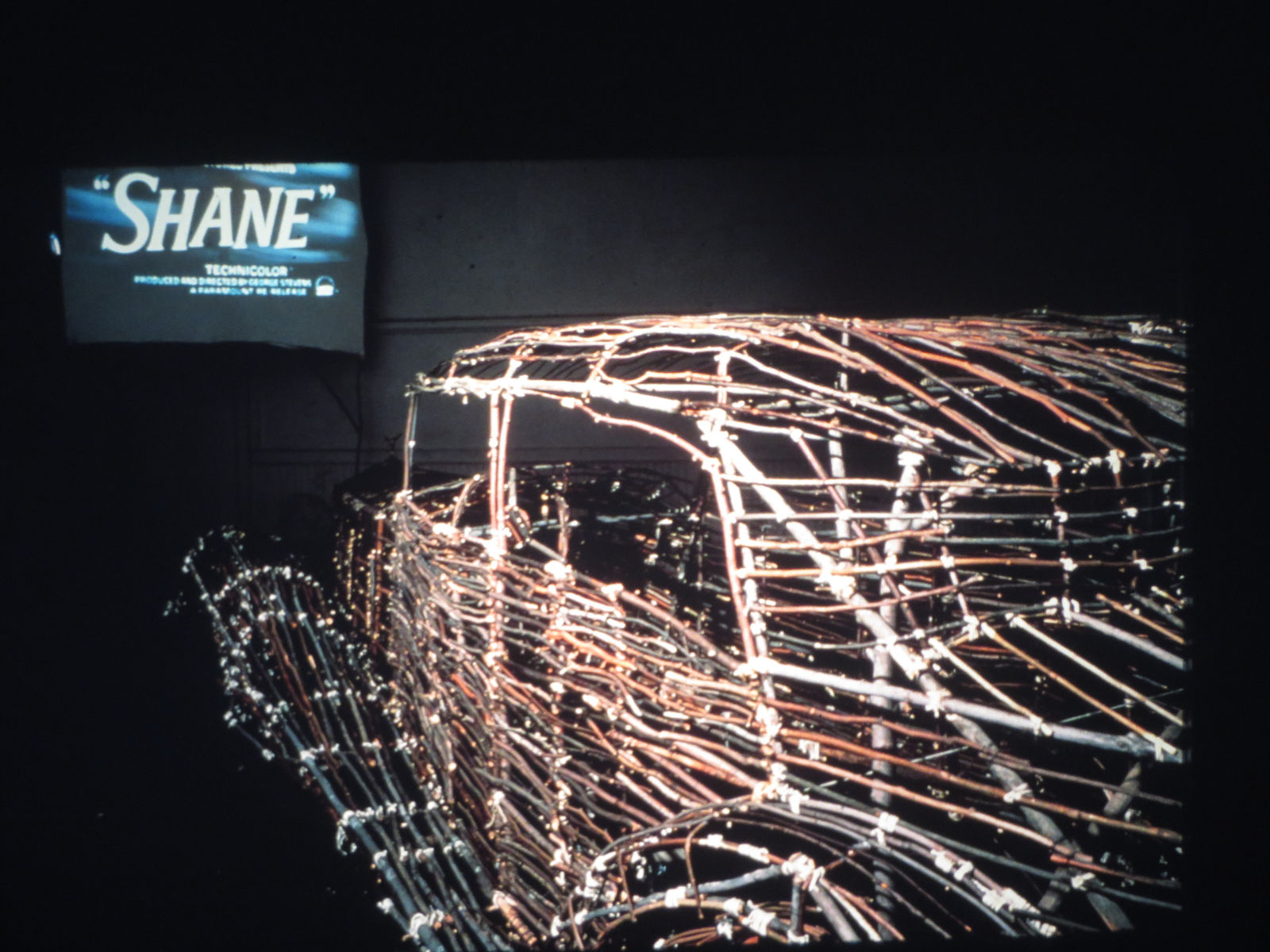 A car made from bamboo and twigs is placed in front of a movie screen to mimic the set-up of a drive in movie theater.