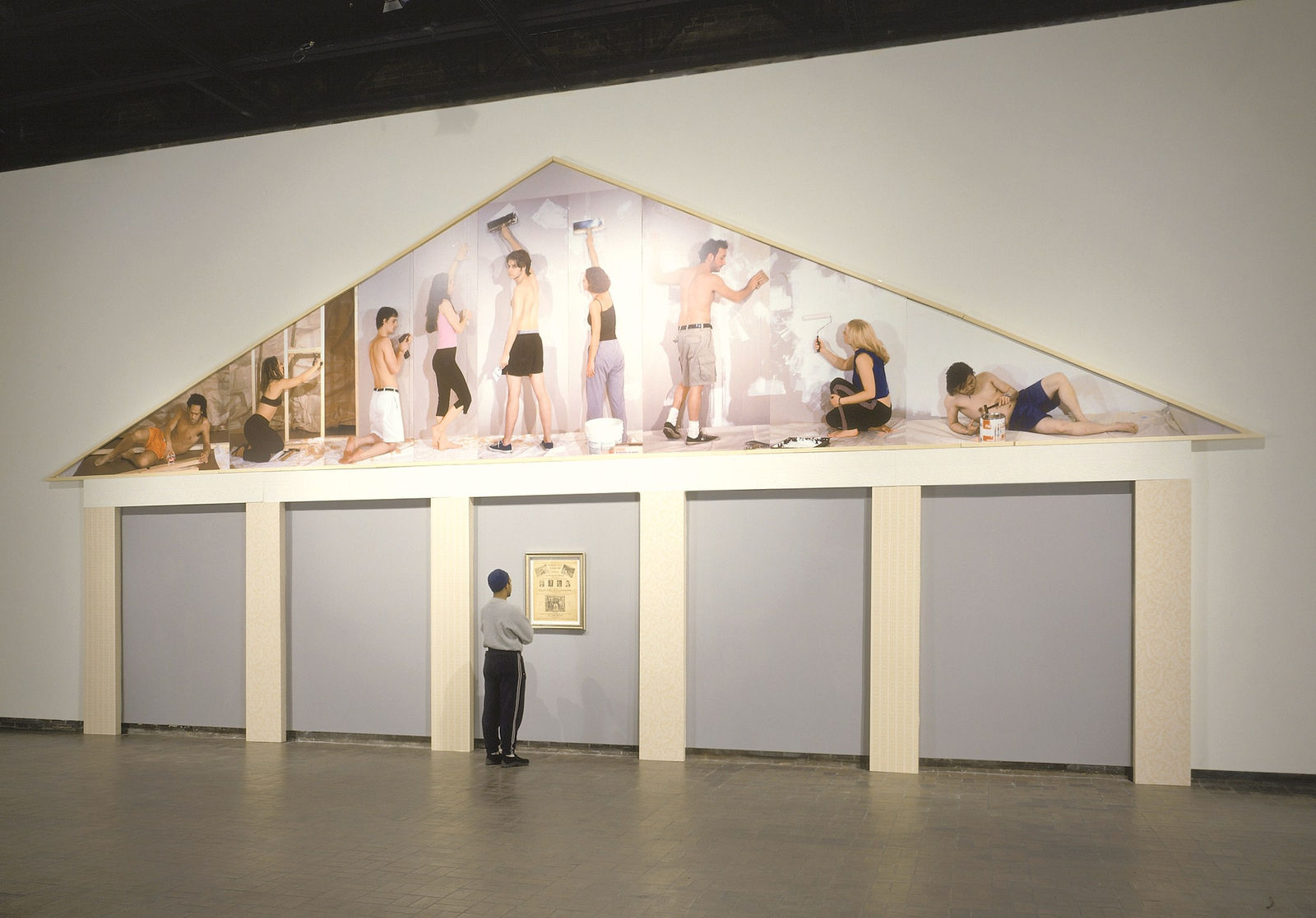 Wooden beams create the outline of a house. 9 figures are painted on the canvas roof. The piece is displayed against a white wall in a museum setting.