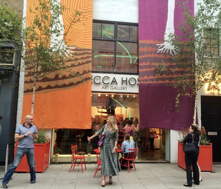 The front of the Rebecca Hossack Art Gallery with two large banners of Aboriginal art hanging from the top of the building. Various people stand inside and in-front of the gallery.