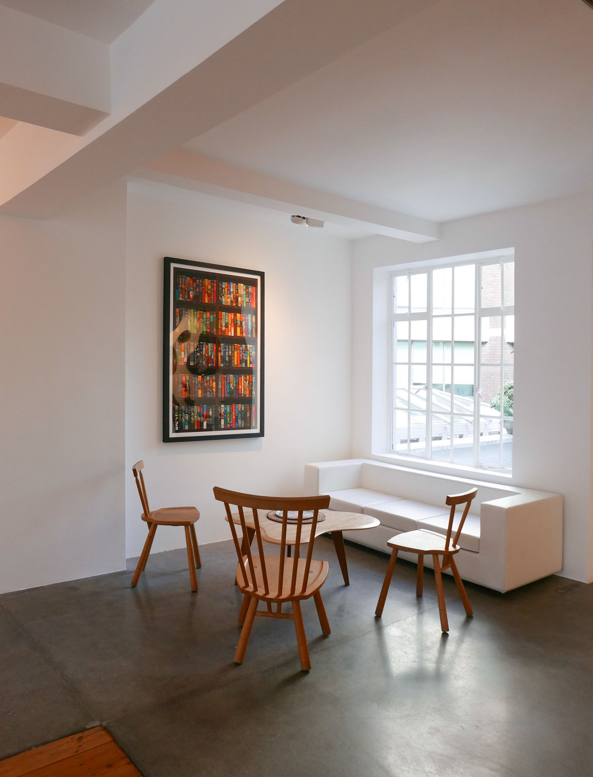 A coffee table in the corner of the Rebecca Hossack Art Gallery, surrounded by three wooden chairs and a white sofa. There is a large, bright window behind the sofa and an artwork by artist Phil Shaw hanging on the wall.