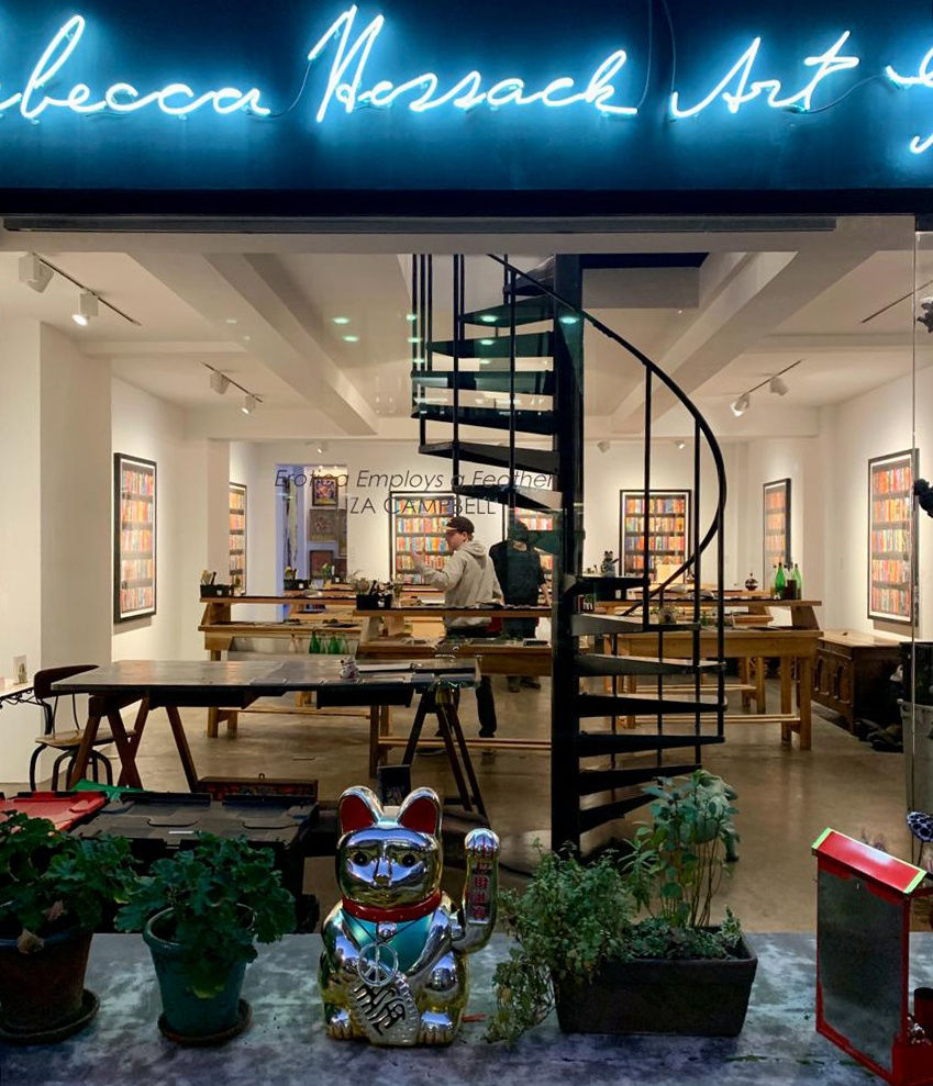 A view through the front window of the Rebecca Hossack Art Gallery. There are tables inside and a person walking around inside. Outside of the window are various plants and money cat on a. table.