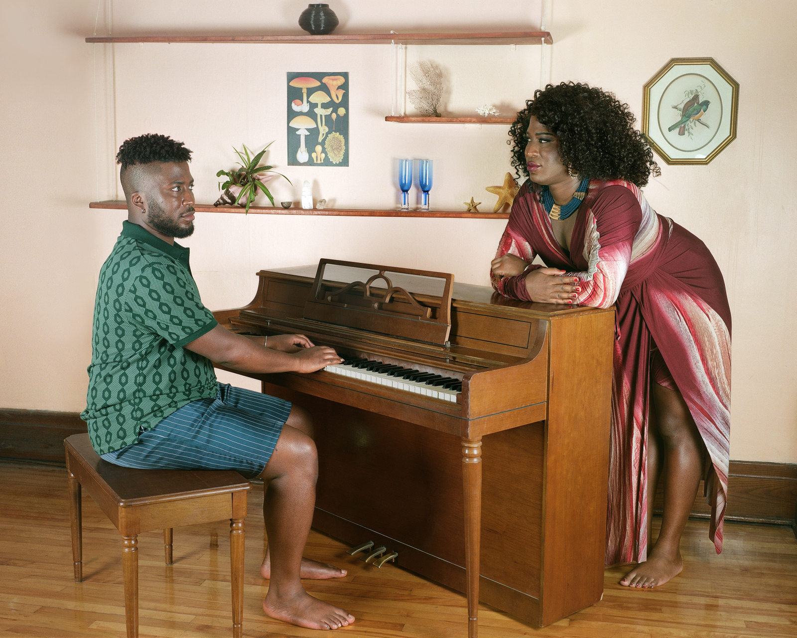 A Black man sits at a piano while looking at a Black woman who is leaning on the piano looking back at him.