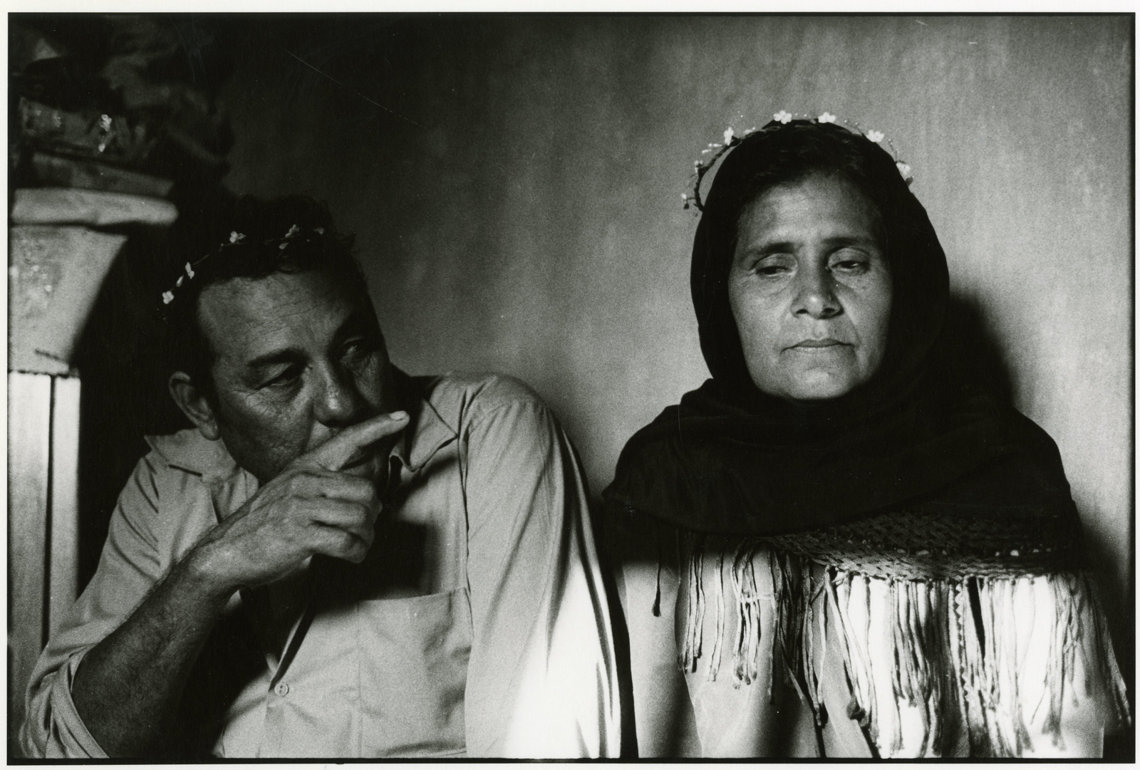 A man and woman are photographed seated next to each other in black and white, with the woman stoic and the man pointing in her direction.