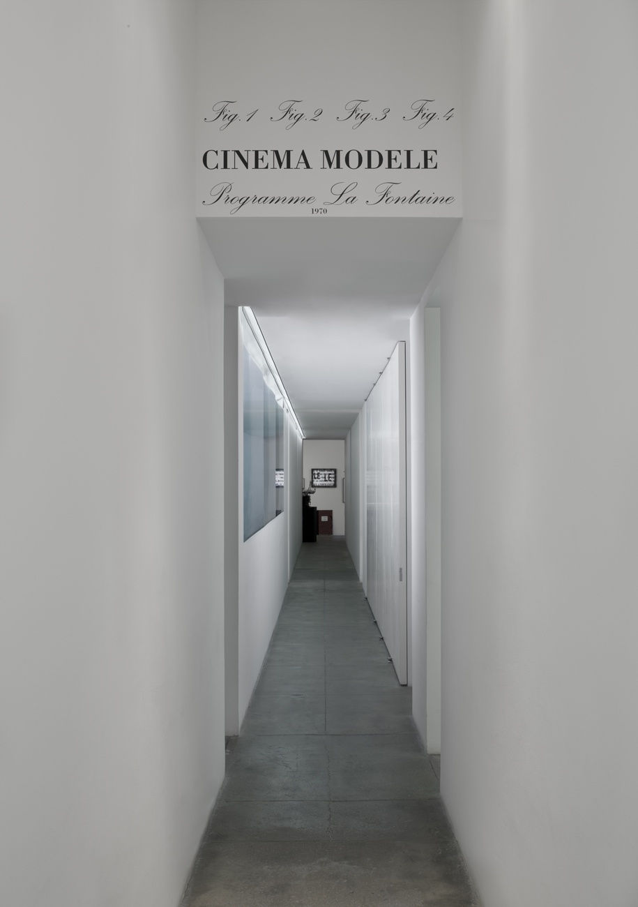 A sign on the ceiling at the start of a long hallway into the exhibition reads, in black cursive: Fig. 1 Fig. 2 Fig. 3 Fig. 4 CINEMA MODELE Programme La Fontaine 1970