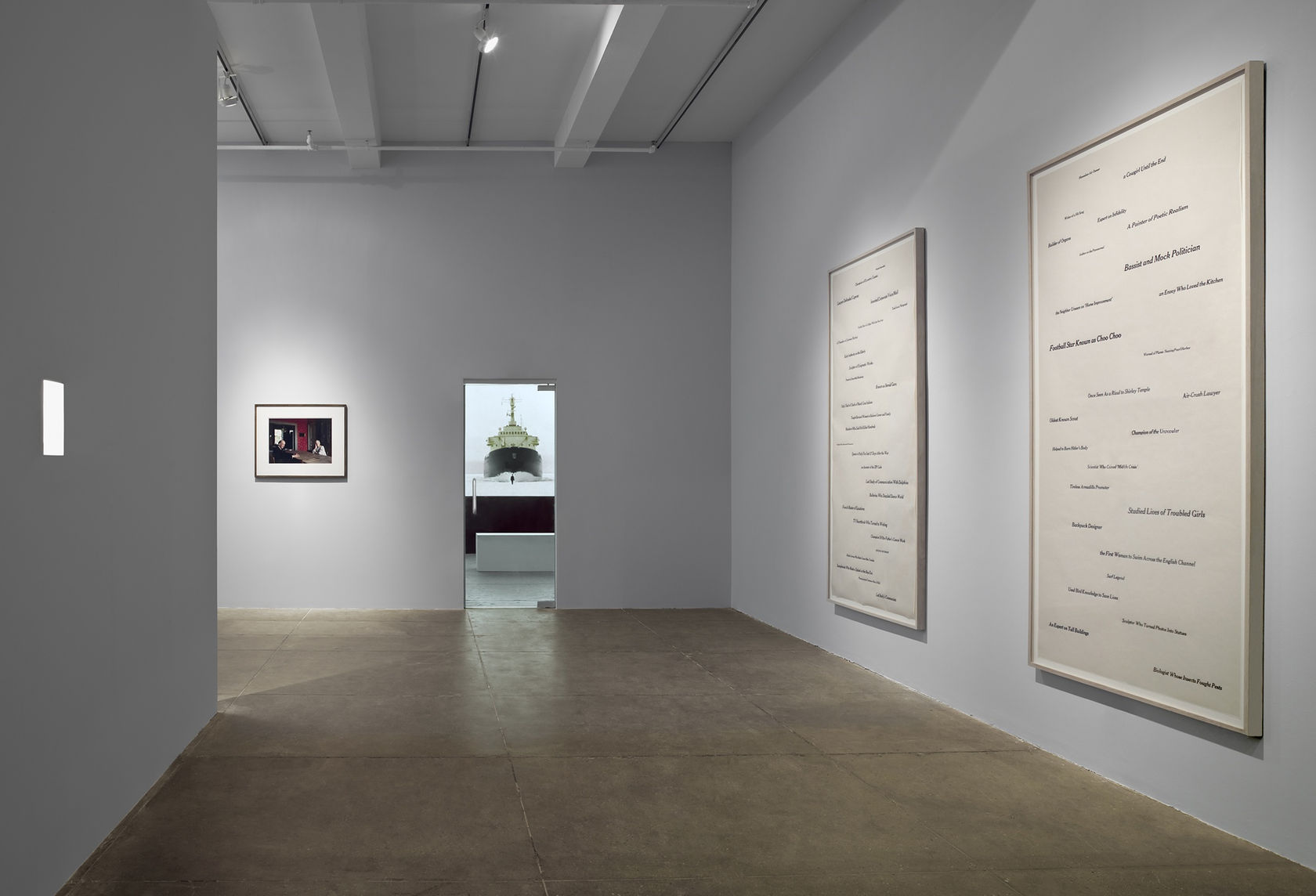 On the left wall there is a square of light. In the center is a model ship visible through a doorway, next to a framed photograph.  On the right wall hang two framed works with text on paper. 