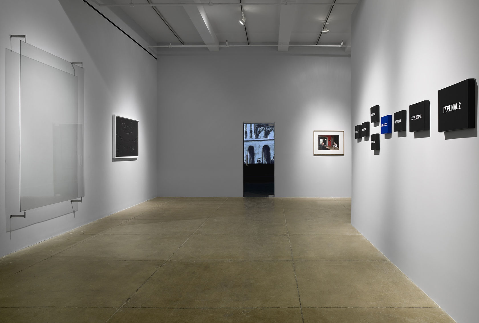 Works of various materials and sizes hang on the walls of a room painted light gray. In the center appears to be a door illuminated by a building lit from within. 