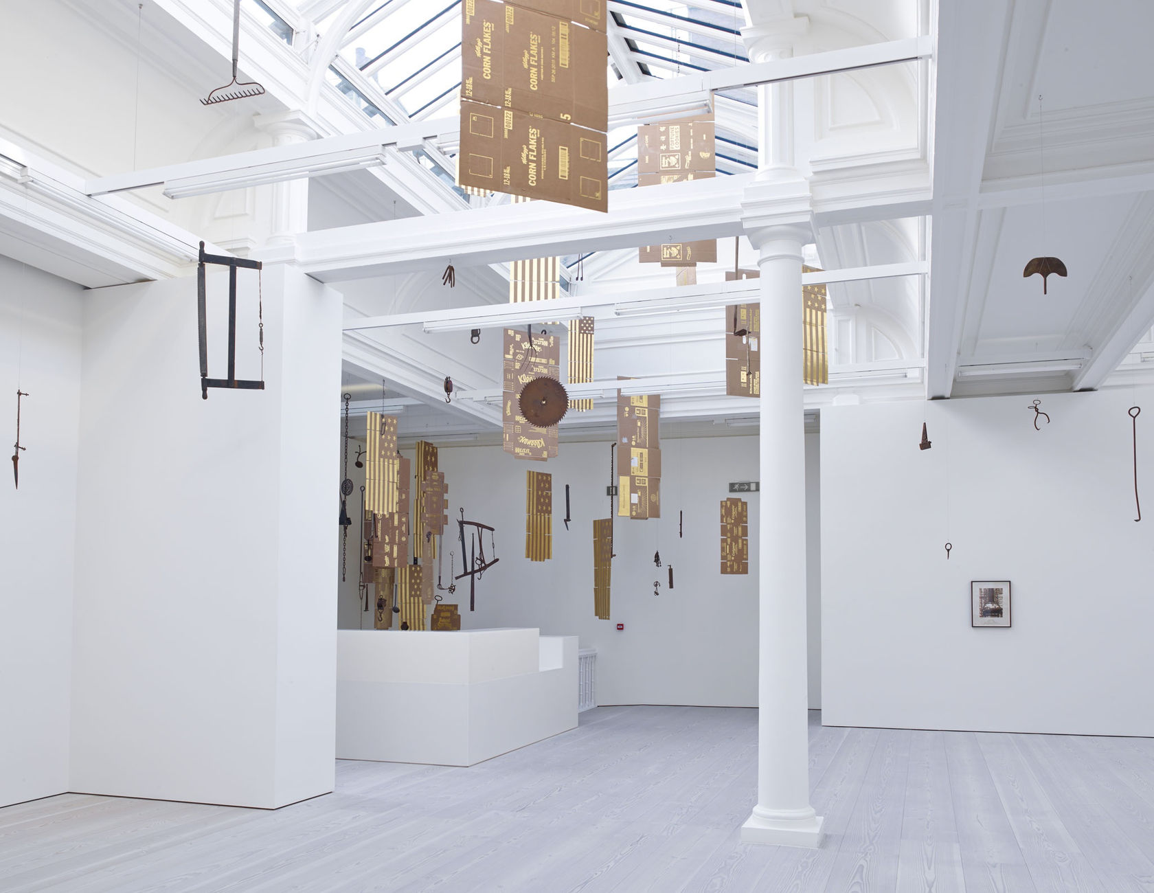In a large white space with a long glass skylight, sculptures hang from the ceiling - most seem to be American flags, with 13 stars, made out of gold painted cardboard. Another is an unfolded Corn Flakes box, made from the same material.