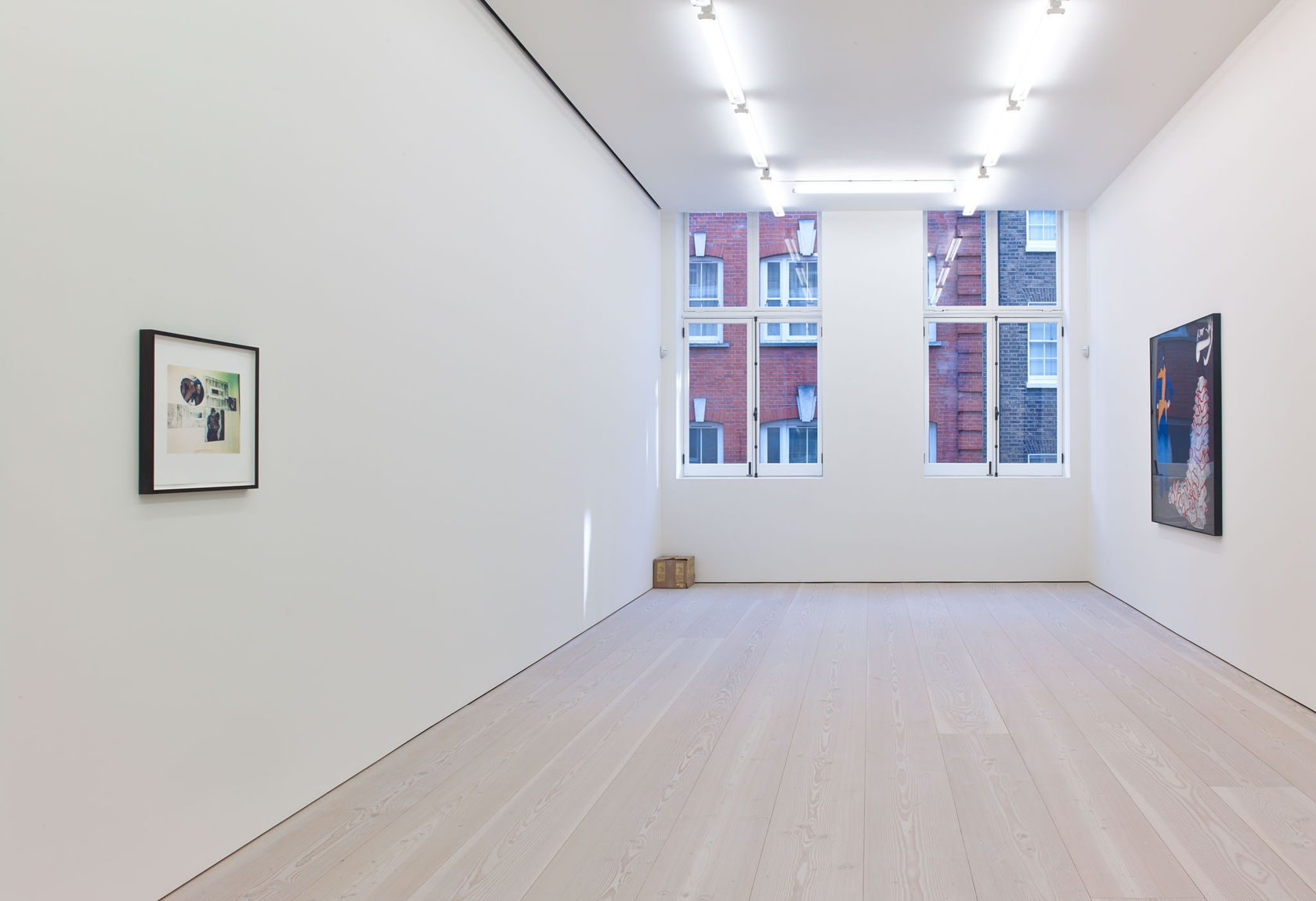 In a white room with windows straight ahead, a small print hangs on the left wall, a large colorful work on the right wall, and a small cardboard box sits in the left corner under the window.