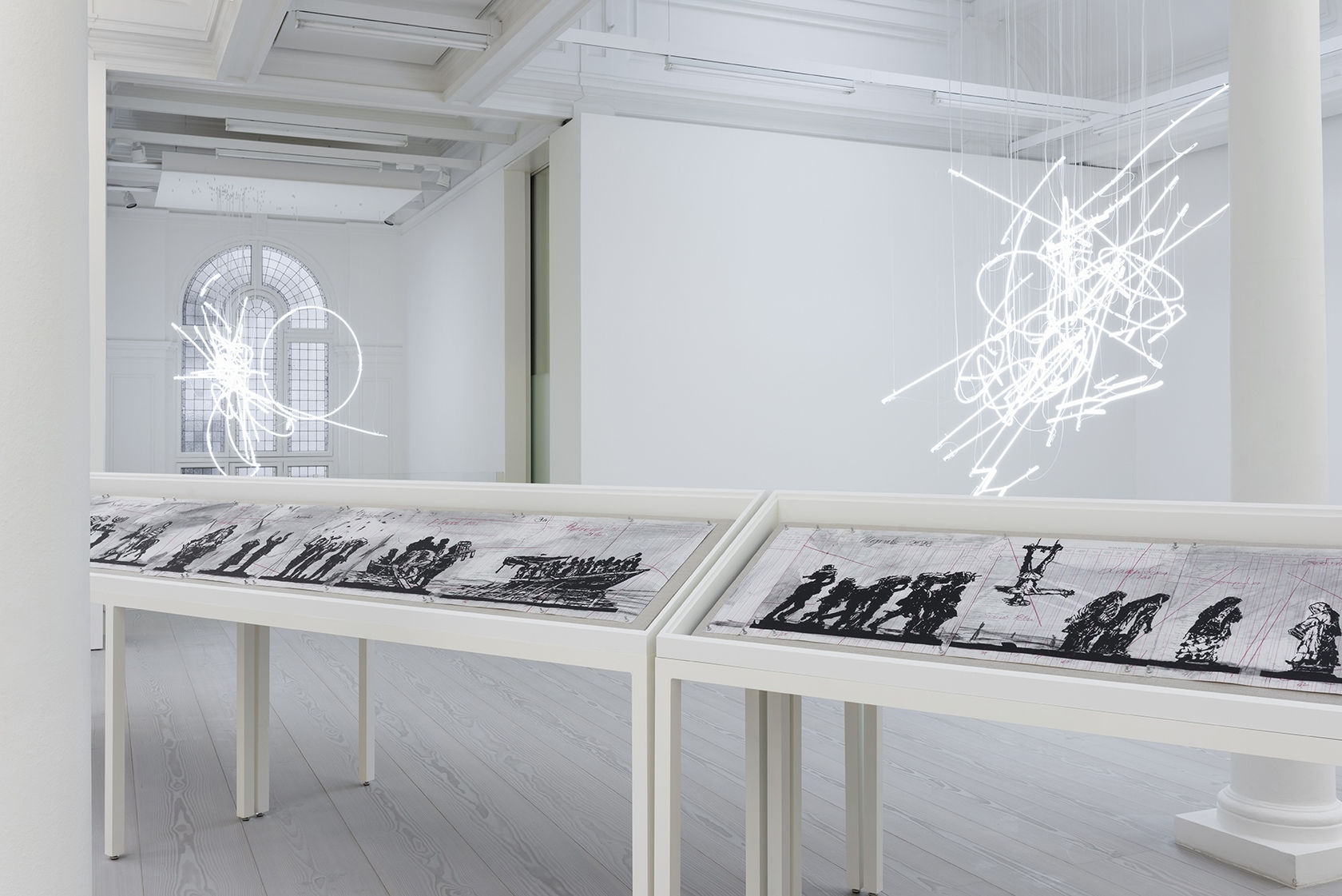 A long vitrine displaying charcoal drawings sits under a neon sculpture hanging from the ceiling.