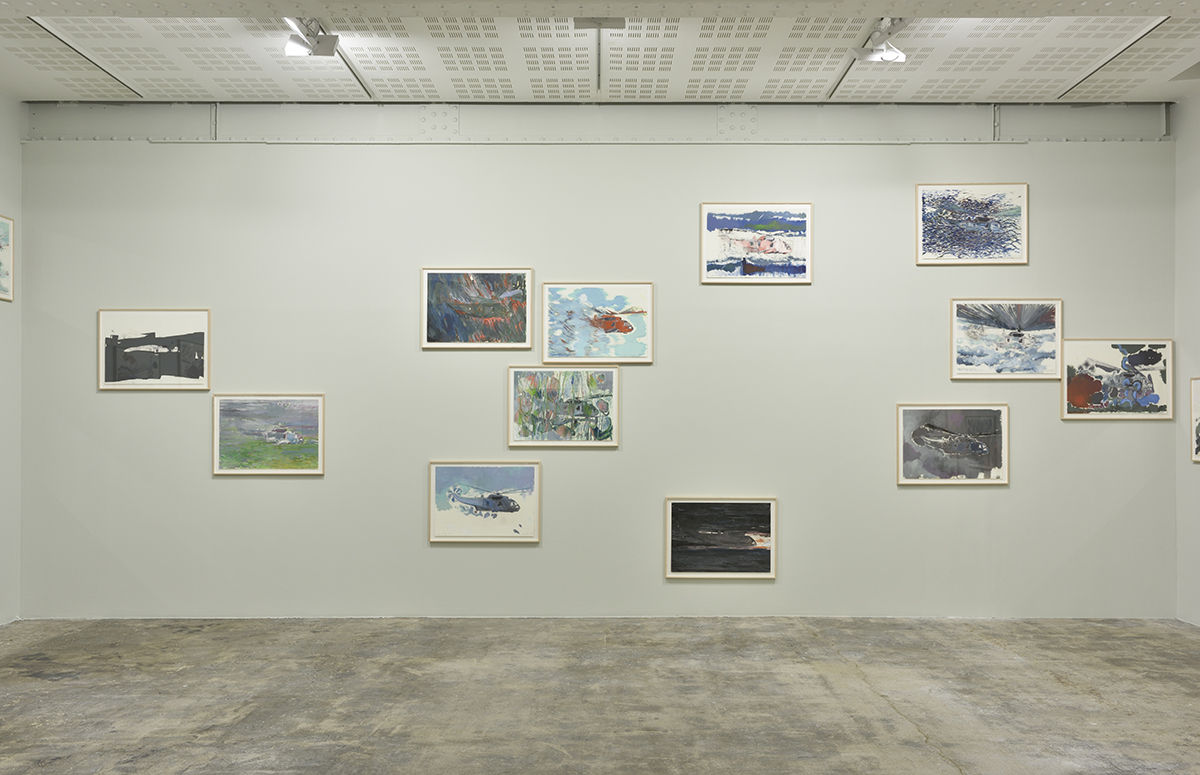 Many smaller, framed paintings of helicopters on paper hung at various heights on a beige gallery wall.