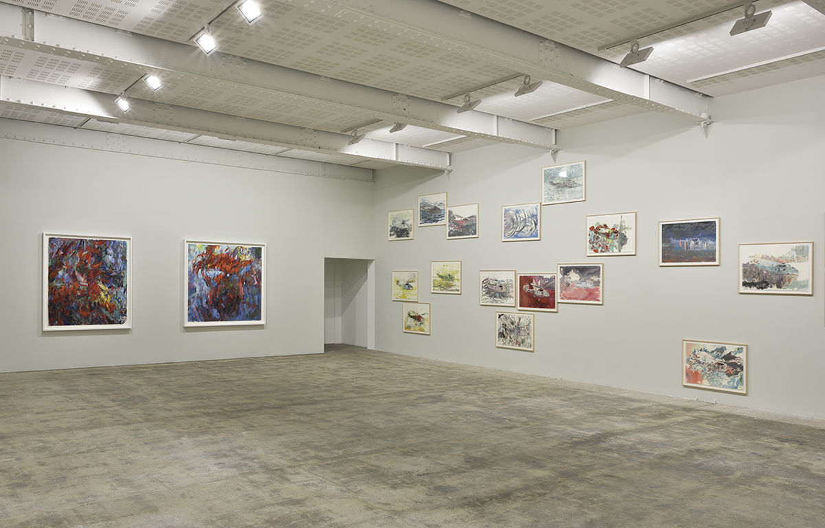 Framed paintings of helicopters on paper and 2 large abstract paintings hang at various heights on a beige wall.