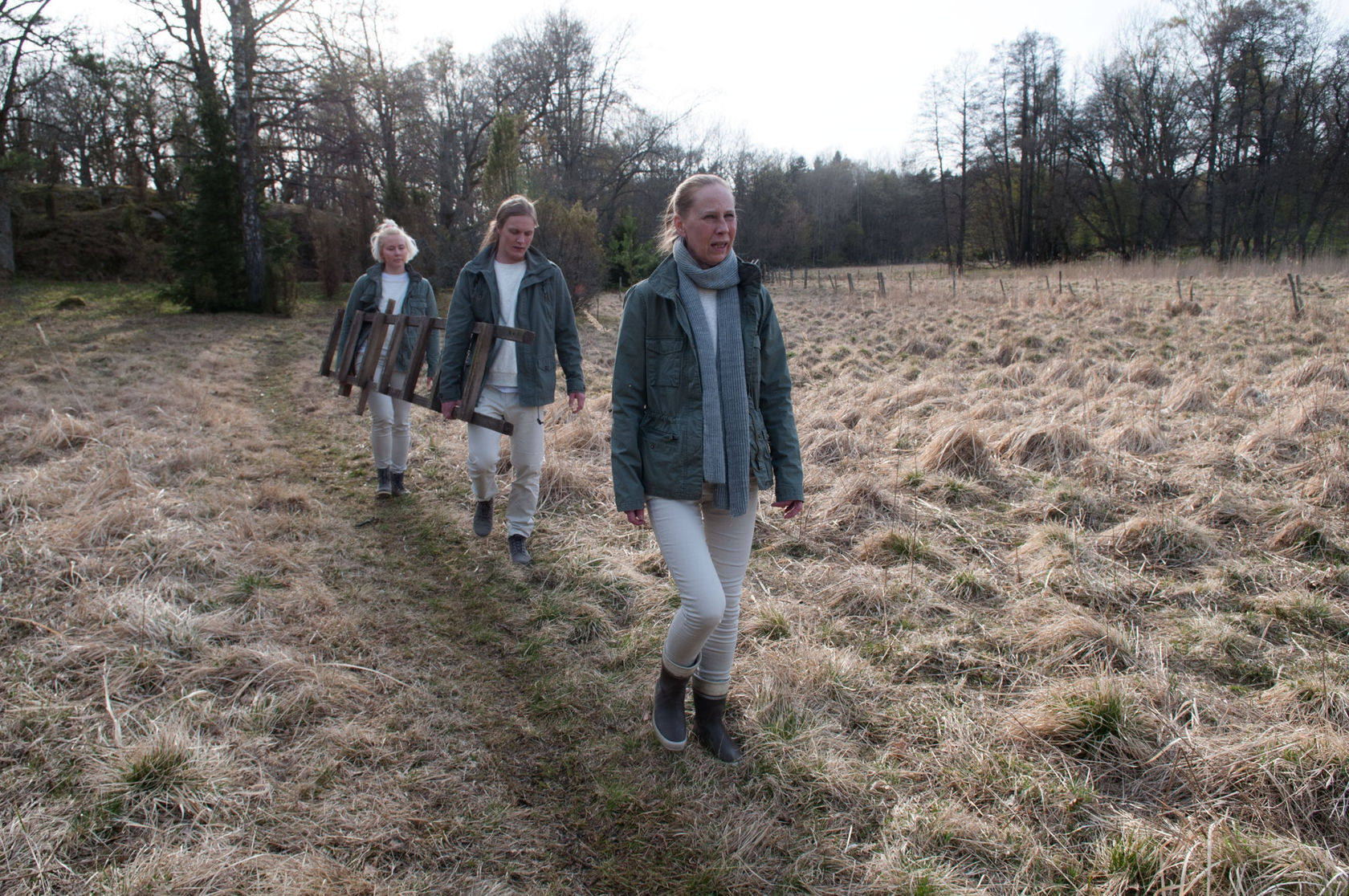 Image of three people in matching green jackets and tan pants walk in a line through a field. Two carry a ladder.