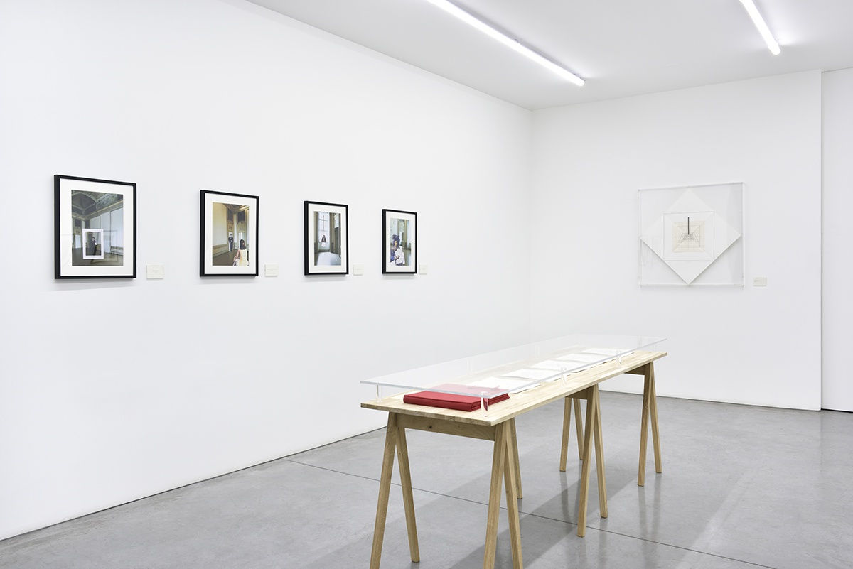 A well lit gallery displays framed works on the wall and a display table in the center of the room.