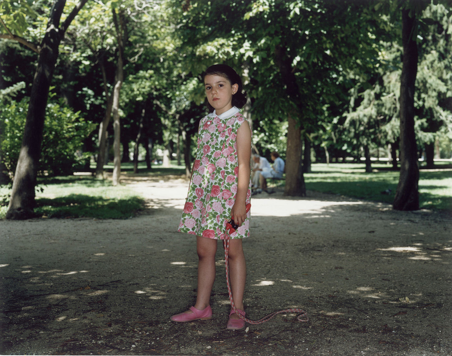 A Rineke Dijkstra photograph: A girl in a floral dress holding a jumprope stands in the center of a tree-lined path in a sunny park. 