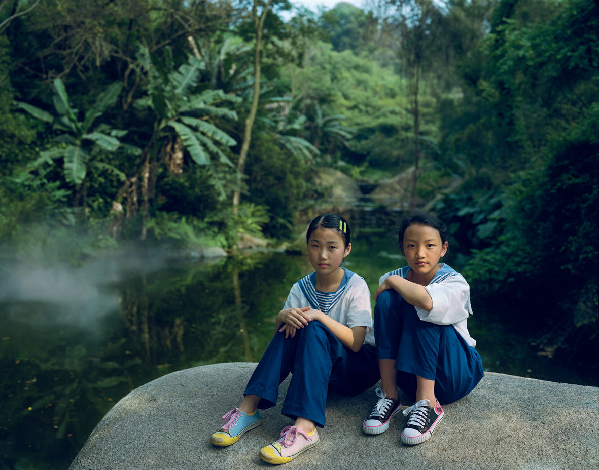 A Rineke Dijkstra photograph: 2 girls in matching uniforms sit on a rock in a lush, green park.