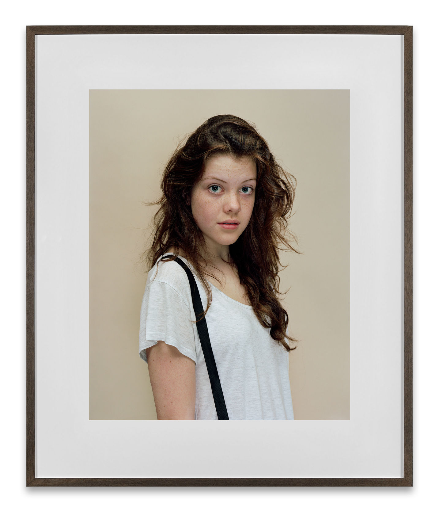 A Rineke Dijkstra portrait of the actress Georgie Henley in a white t-shirt and suspenders in front of a neutral background. 
