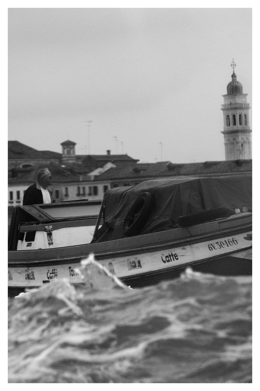 Black and white photograph of a man on a boat delivering coffie in Venice, Italy.