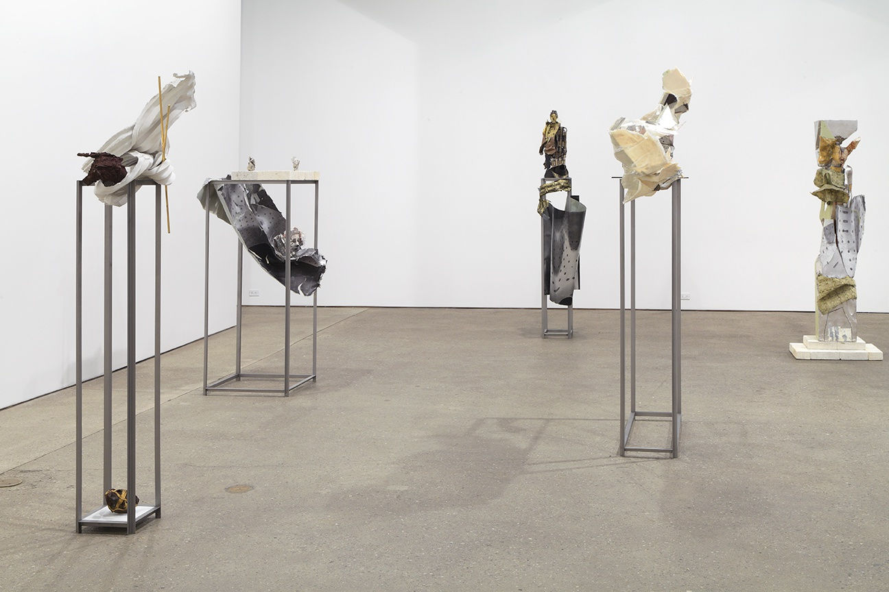 Installation view of Matthew Monahan's sixth solo show at Anton Kern Gallery, featuring sixteen sculptures, a set of drawings, two wall-mounted masks, and a single large figure in the back room. A few sculptures.