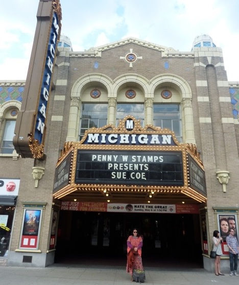 The artist stands in front of a theater. The theater's billboard announces that Sue Coe will be giving the lecture for the Penny Stamps Distinguished Speaker Series