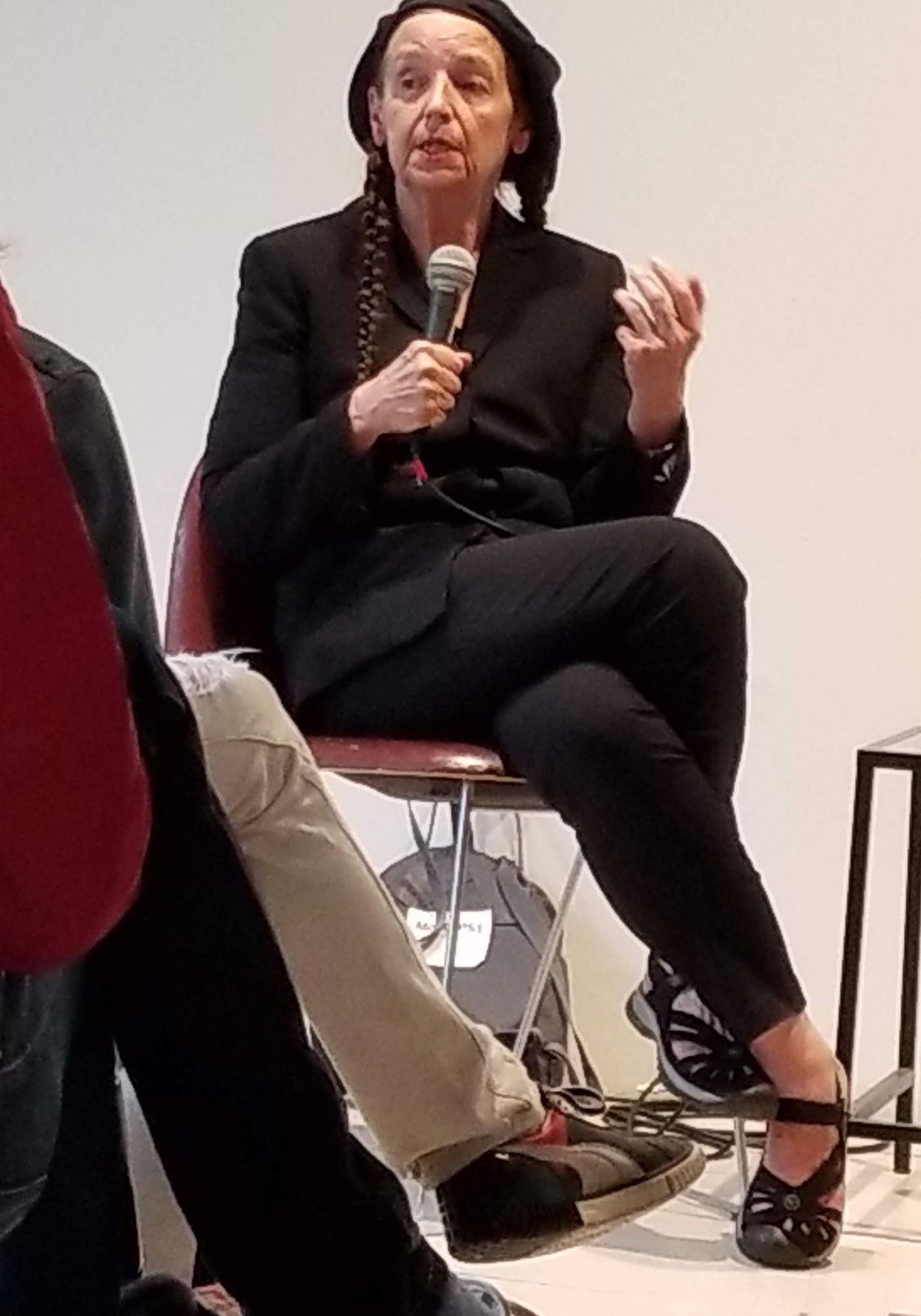 The artist Sue Coe answers a question into a microphone during a chat with the curator Peter Eleey at the opening of her exhibition "Graphic Resistance" at MoMA PS1. She wears a black beret and an all black outfit.
