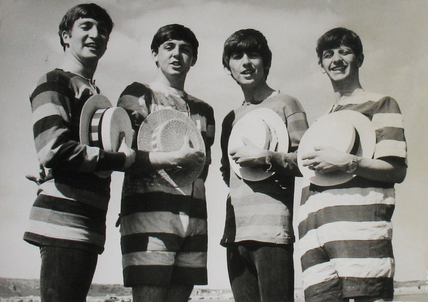 Black and white photograph of the Beatles on the beach in stripped shirts holding straw hats