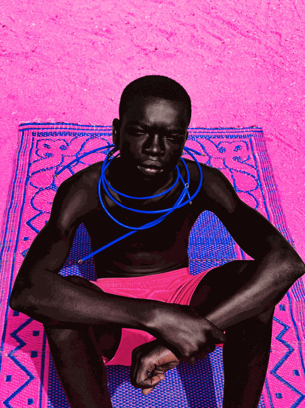 Three-quarter length portrait, seen from slightly above, of a shirtless young Black man wearing bright pink shorts, seated on a blue and pink patterned rug the ground, his elbows on his knees, against a bright pink background.