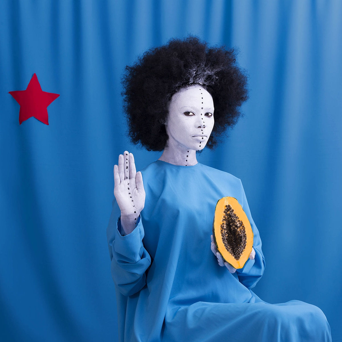 A seated woman with a white-painted face, black hair, and wearing a blue nun-like garment, holds her right hand up as if taking an oath, her palm also painted white, while holding a halved fruit in her left hand, its seeds visible to the viewer.