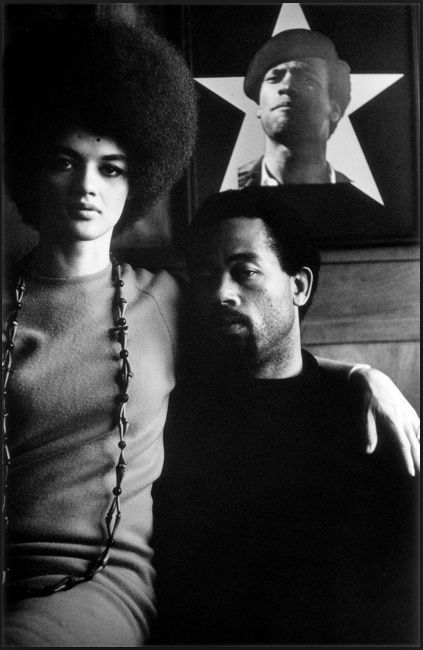 Kathleen and Eldridge Cleaver in black and white, seated in front of a poster of Huey Newton, whose head emerges from a white star background.