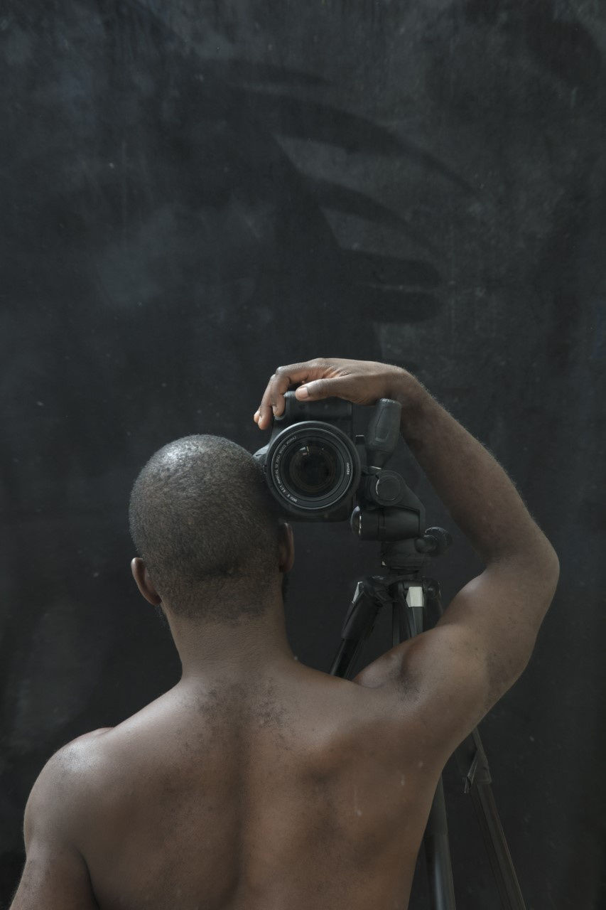 Half-length portrait of a shirtless Black man, seen from behind, with his right arm raised holding a camera against his head, its lens aimed at the viewer.