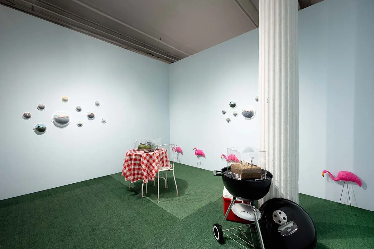 Abigail Goldman installation at Outsider Art Fair. Picturesque suburban backyard with white picket fence in forground, red checkered tablecloth bistro table and grill / cooler visible. Abigail's work hangs in clusters on walls and on top of grill and tabl