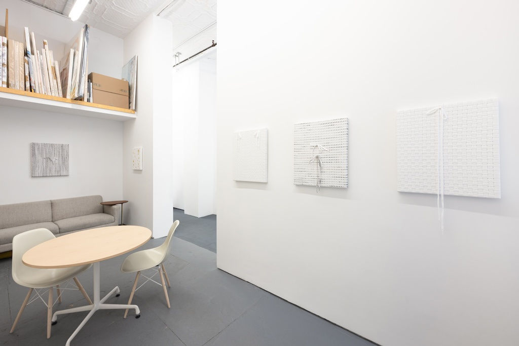 Installation of Didi Rojas exhibition - white shoelace paintings on wall, round table and white chairs to the left