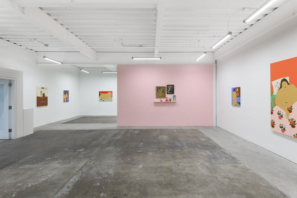 installation of Gina M. Contreras exhibition - pink wall to center with small sculptures and religious paintings, white wall along back and right side. Paintings of nude female figures