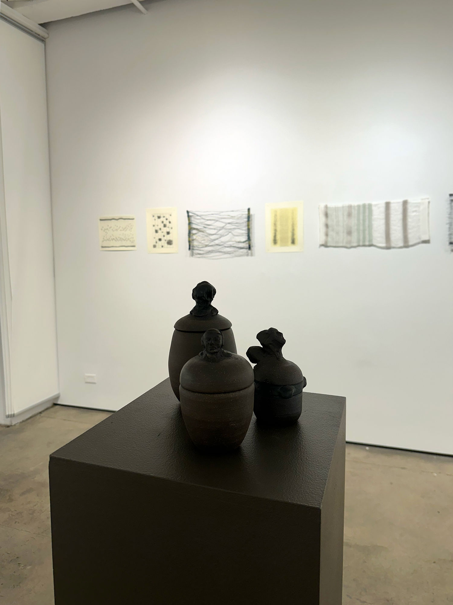 photo of an installation with three small urns on a pedestal. behind them on the wall are a series of small works.
