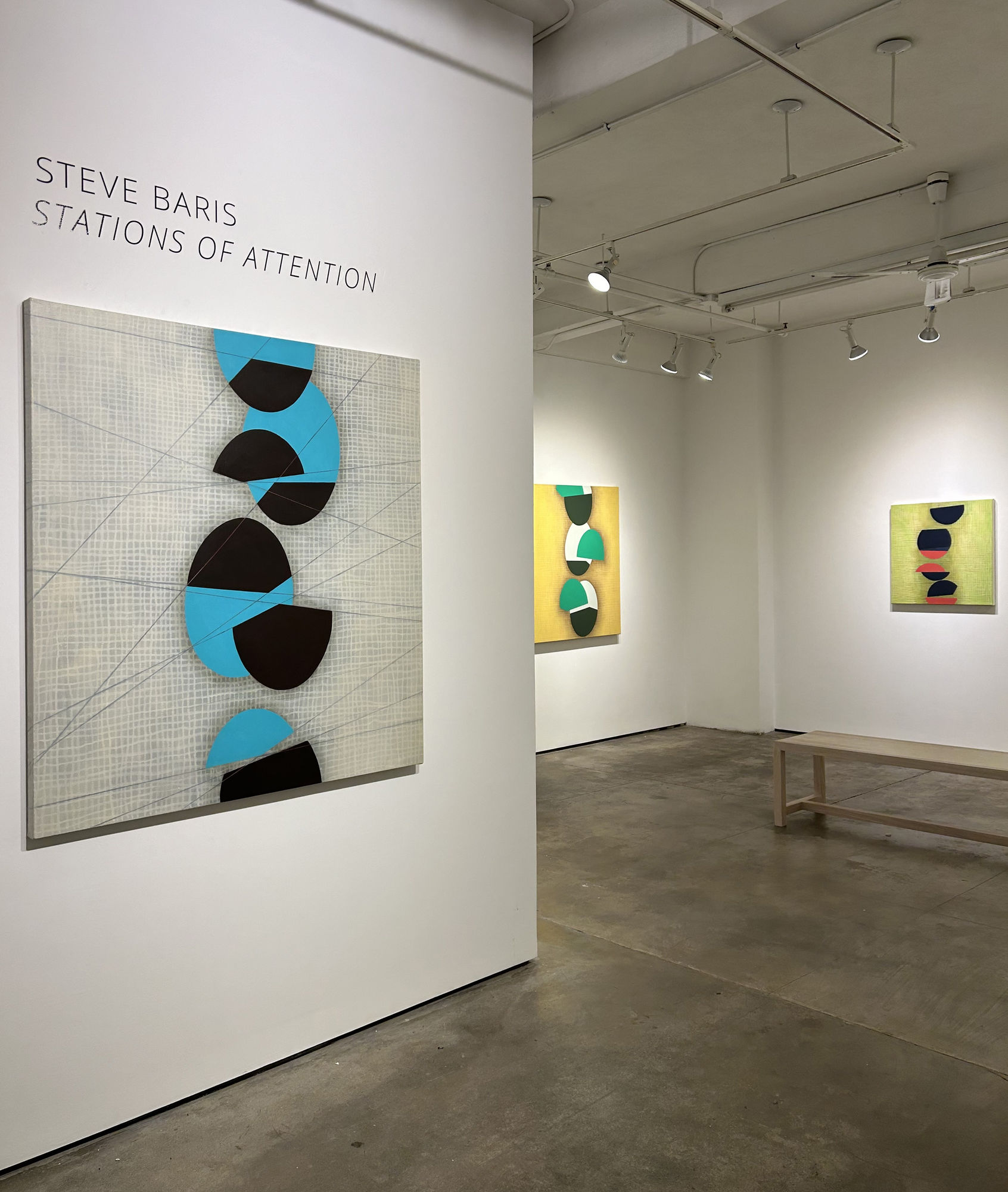 Photo of a Steve Baris exhibition, called Stations of Attention. There are three paintings visible on the walls. Each painting has a low contrast painted grid in the background, with a series of circular shapes going up the center, which appear to be bise