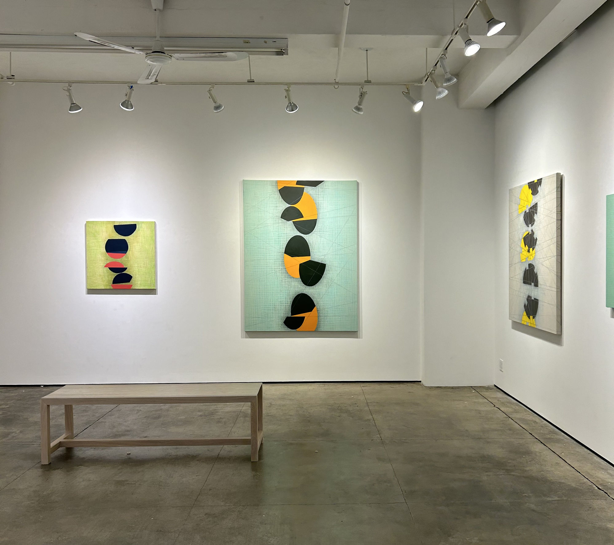 Photo of a painting exhibition, with three works visible, and a bench in the center of the room.  Each painting has a low contrast painted grid in the background, with a series of circular shapes going up the center, which appear to be bisected at various