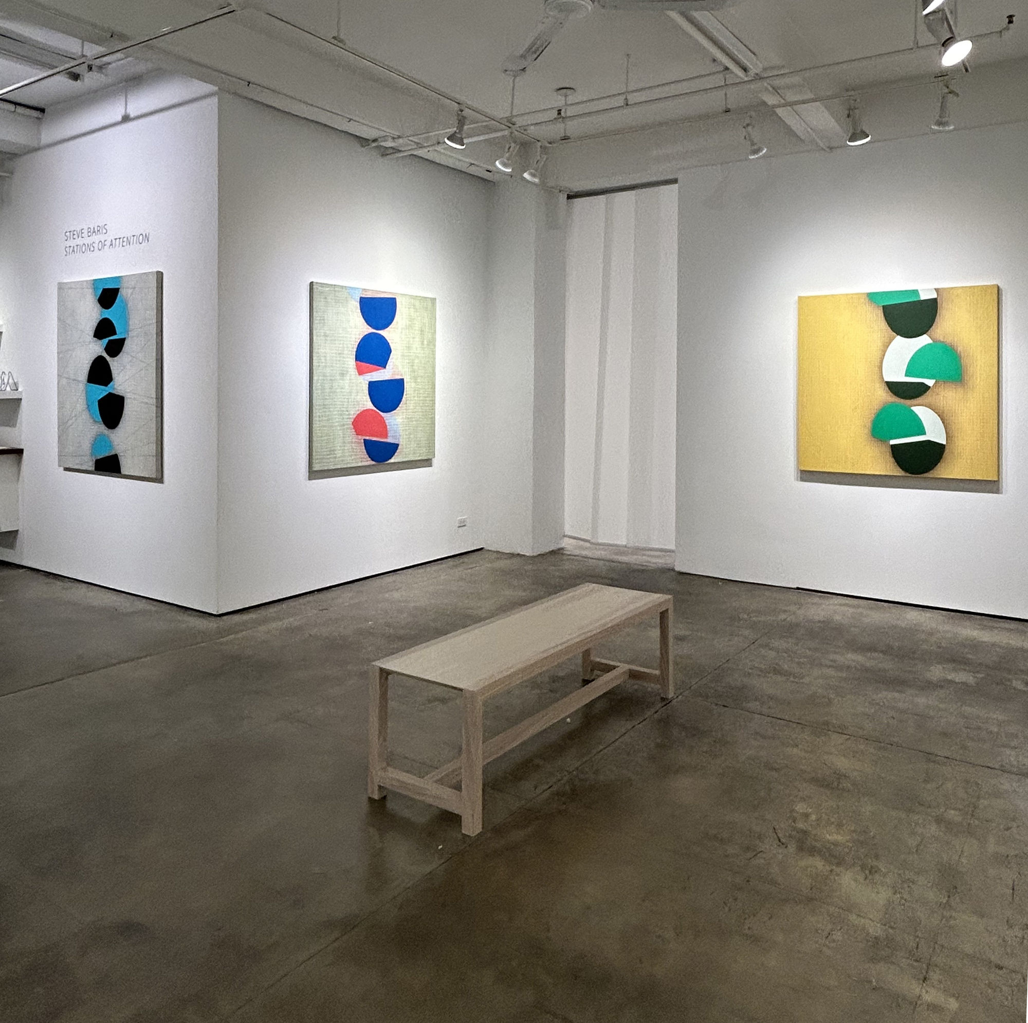Photo of a Steve Baris exhibition, called Stations of Attention. There are three paintings visible on the walls. Each painting has a low contrast painted grid in the background, with a series of circular shapes going up the center, which appear to be bise