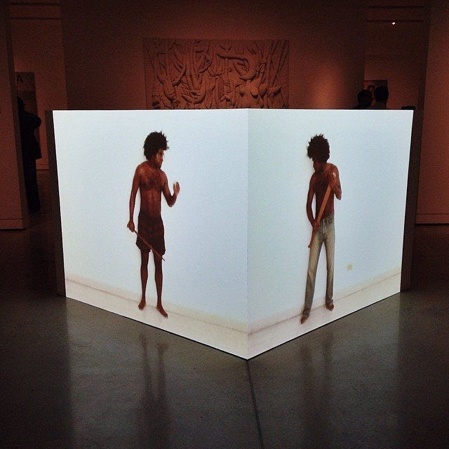 A white cube sits in the centre of the room. Two figures are projected onto different walls of the cube, facing each other.