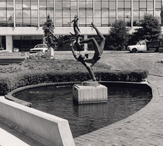 Black and white photograph of a sculpture in the middle of a small pond. The sculpture depicts a figure holding itself up on one hand, while balancing smaller figures on its foot in mid-air.
