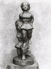 Black and white photo of a wooden sculpture. The sculpture depicts two female figures with one figure being lifted on the shoulder of the other.